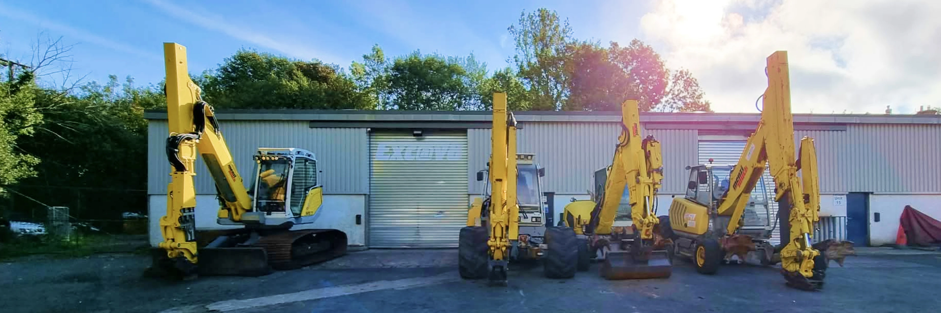 Exc@v8's workshop and yard in Kendal, Cumbria, with a line of Menzi Muck walking excavators parked outside the servicing bay, the Exc@v8 sign visible on the shutter door