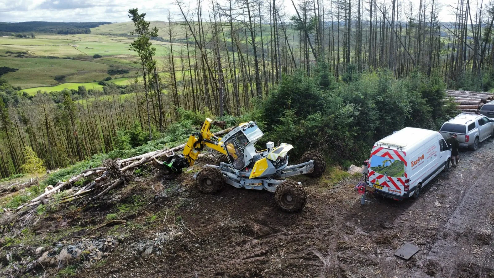 An Exc@v8 walking excavator working a forestry site, with the team's liveried van and support vehicle parked on the track