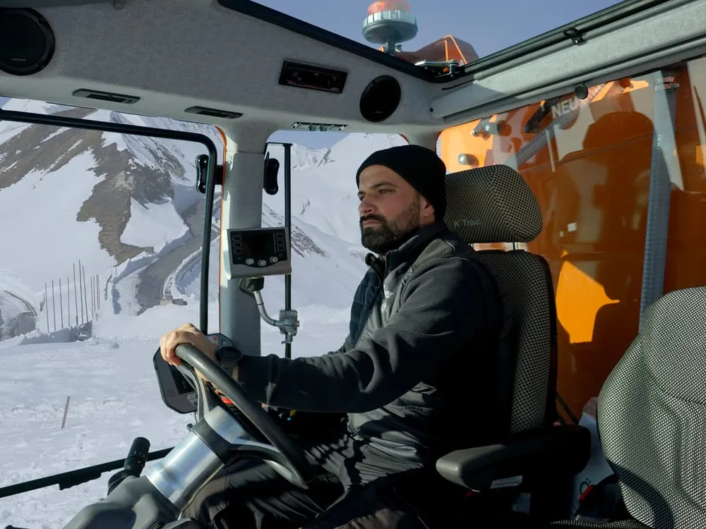 An operator in the K Trac’s cab, with wide windows onto a snowy mountain road